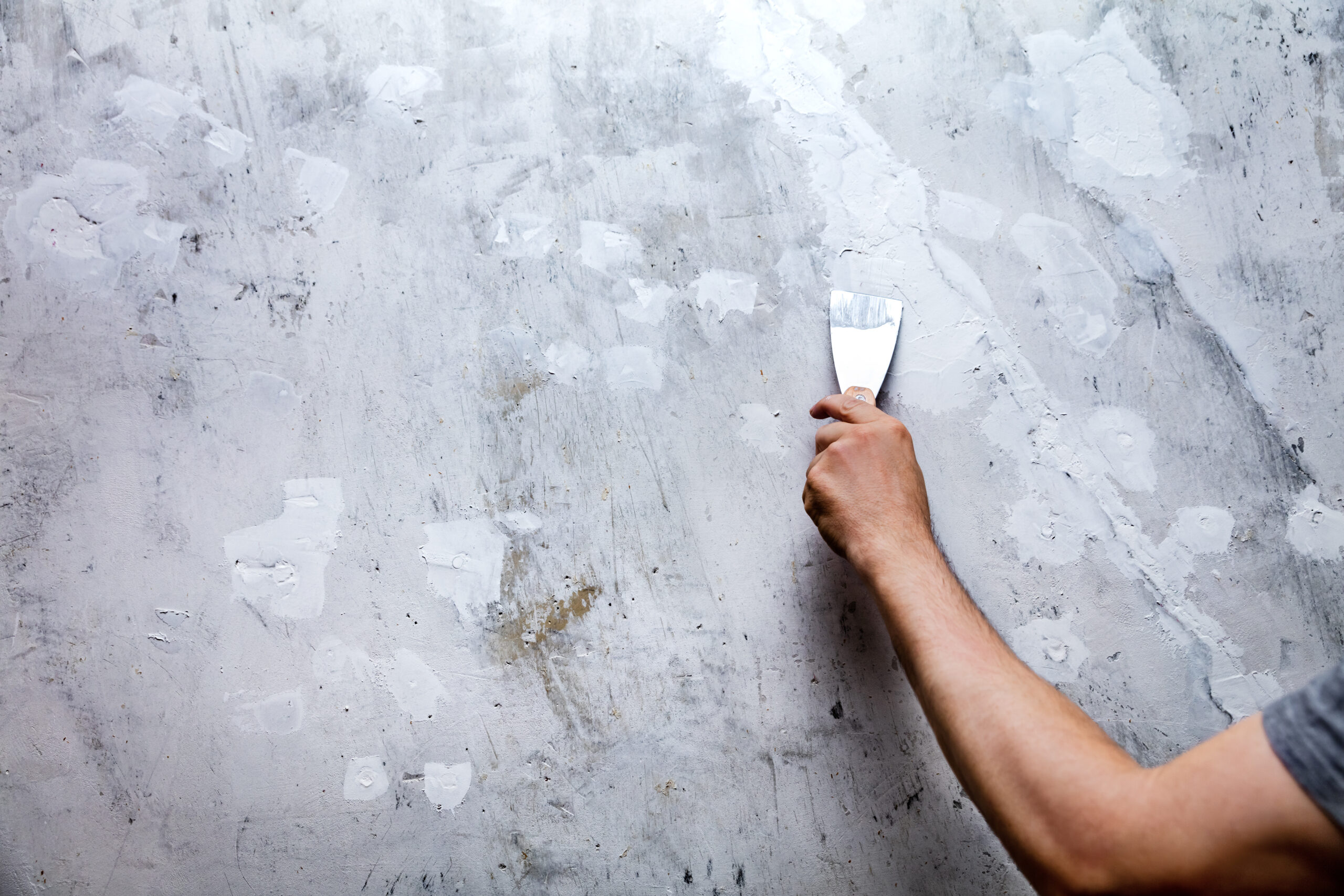 Ein Handwerker trägt mit einem Spachtel eine weiße Beschichtung auf eine graue Wand auf.