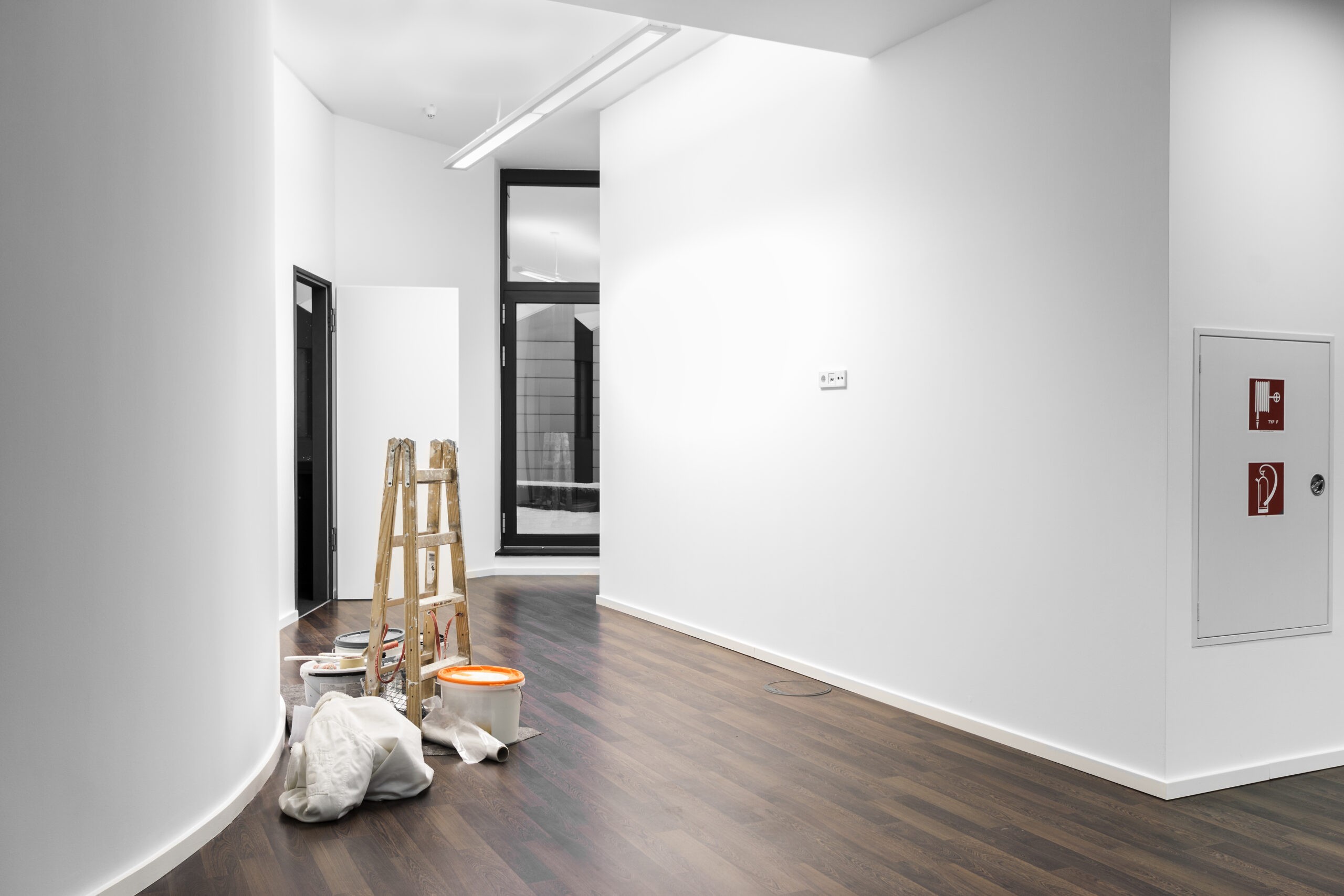 Ein heller Flur mit dunklem Holzboden, einer Holzleiter und Farbeimern während Renovierungsarbeiten vor einer weißen Wand mit einem Wandschrank.